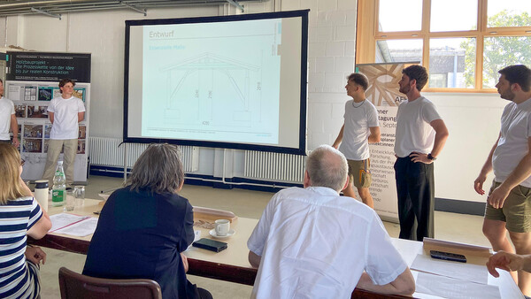 Die Zeichnung einer Holzkonstruktion (mit der Aufschrift "Entwurf") wird auf eine Leinwand geworfen, rechts und links davon stehen junge Menschen in lässiger Kleidung. Am vorderen Bildrand, nahe dem Betrachter, sitzen Personen an langen Tischen mit Dokumenten (und Getränken) vor sich. Offenbar hören sie einer Präsentation zu oder bewerten sie. Ganz links steht ein Rollup mit der Aufschrift "Holzbauprojekt - Die Prozesskette von der Idee bis zur realen Konstruktion".