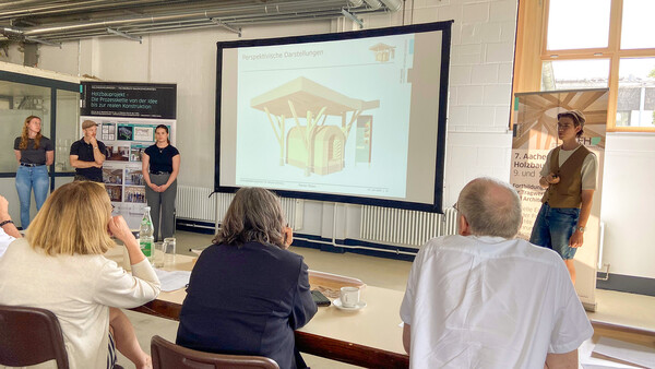 Eine Leinwand wird mit der Zeichnung eines Bauprojekts bestrahlt: Eine Art Tunnel mit eckigem Boden und runder Oberseite, darüber ein hölzernes Schutzdach. Links und rechts der Leinwand stehen einzelne Menschen, offenbar Vortragende. Im Vordergrund sitzen Personen an einem langen Tisch, ausgerüstet mit Kaffee, Wasser und Schreibkram. Links weist ein Rollup darauf hin, dass es sich um "Holzbauprojekt" handelt, rechts weist ein Rollup auf die 7. Aachener Holzbautagung hin.