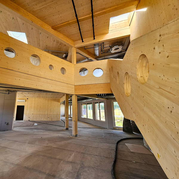 Das Innere eines Gebäudes, das bis auf den Betonboden komplett aus Holz besteht. Zu sehen sind mehrere Stockwerke mit halb offenen Holzwänden mit kreisförmigen Öffnungen darin, und hölzerne Säulen, die die Stockwerke stützen.