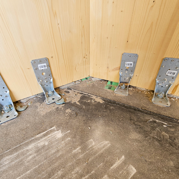 Vier Holzverbinder in der Ecke eines Raumes, dessen Wände aus Holz und dessen Boden aus Beton bestehen. Jeweils zwei Winkelverbinder links und rechts der Ecke.