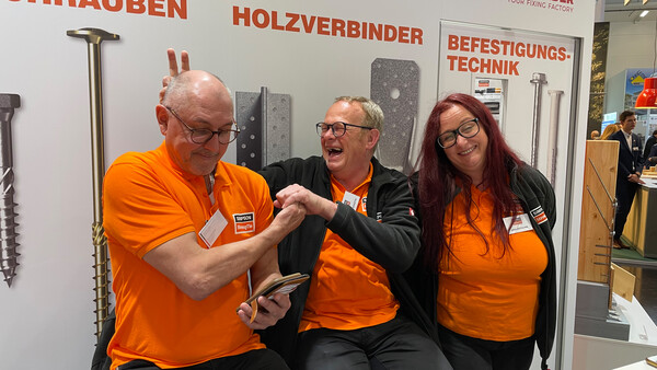 Drei Mitarbeiter von Simpson Strong-Tie sitzen vor einer Messewand mit Produktpräsentation, scherzen miteinander und lachen