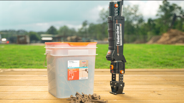 Eine hölzerne Fläche, die ein Boden oder eine Tischplatte sein könnte, dahinter eine grüne Wiese und verschwommen Gebäude, Bäume und Hügel. Auf der Holzfläche ist ein Timber Drive abgesetzt, mit dem flchen Anschlag für Holz-Holz-Anschlüsse auf dem Holz. Daneben steht ein eckiger Behälter aus semitransparentem Kunststoff, der Schrauben enthält - ein paar dieser Schrauben liegen vor dem Behälter. Die Schrauben sind bräunlich und haben sechseckige Löcher.