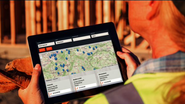 Ein Bauarbeiter in gelber Warnweste kommt von rechts ins Bild, in den Händen hält er einen Tablet-Computer. Das Tablet ist der Fokus des Bildes, auf dem Bildschirm ist eine Website mit Kartenansicht und Suchfunktion abgebildet. Die Karte ist voller blauer Pfeile, darunter sind drei Adressen nebeneinander angeordnet - offenbar Suchergebnisse. Im Hintergrund des Bildes ist leicht verschwommen eine Baustelle mit vielen Holz-Bauelementen zu sehen.
