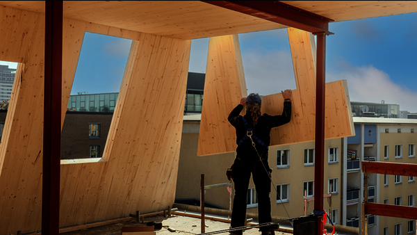 Eine Baustelle in einem höheren Stockwerk, auf der vorgefertigte Holzelemente mit Löchern für Türen und Fenster montiert werden. Eines der Elemente ist in der Schwebe - vermutlich an einem Kran - und wird von einem Bauarbeiter in Position gebracht. Der Bauarbeiter trägt schwarze Kluft mit einem Harnisch an einer Sicherheitsleine, sowie einen Blauen Sturzhelm, unter dem ein Bündel brauner, lockiger Haare hervorschaut. Im Hintergrund sind die Häuser und Teile der Skyline einer größeren Stadt zu sehen.