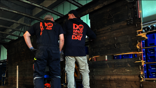 Zwei Männer stehen mit dem Rücken zur Kamera. Sie stehen in einem Waggon aus Holz.