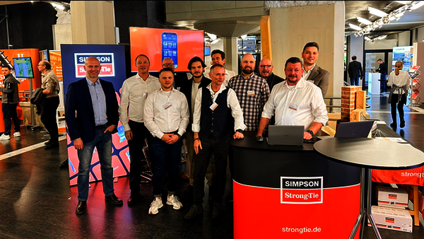 Ein Messestand mit einer rot-schwarzen Theke, einem Stehtisch, einem beleuchteten Display und einem Monitor auf einer hohen Stele. Eine zehnköpfige Mannschaft ist vor dem Stand aufgebaut und lächelt in die Kamera. Alle sind unterschiedlich gekleidet, haben aber Namensschilder am Hemd.