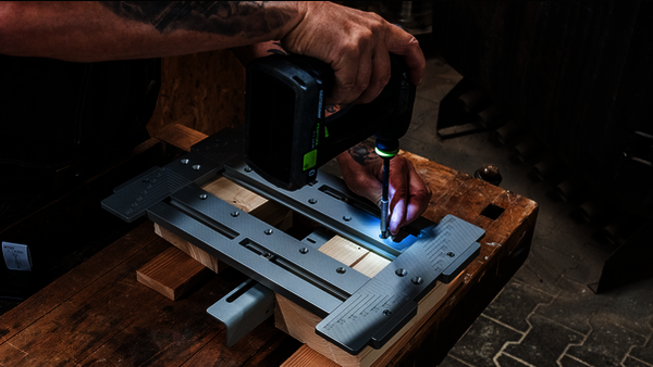 Die Fräs- und Montageschablone MIMO315 von Simpson Strong-Tie in einer Holzbauwerkstatt während der Montage eines Verbinders. Links ist eine Person zu sehen, die einen Akkuschrauber in der Hand hält und damit die Schablone auf einigen auf der Werkbank bereitliegenden Holzbauelementen fixiert.