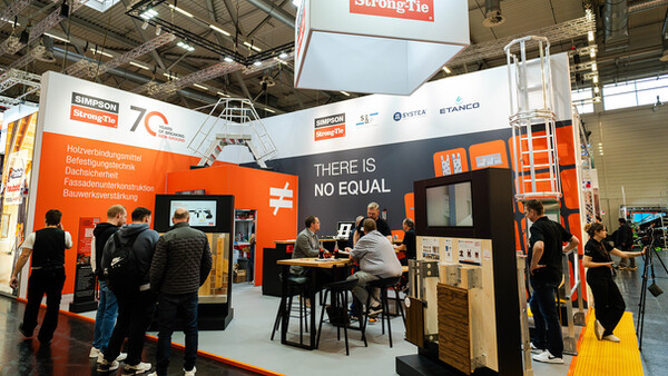 Ein Foto von einem Messestand auf der Dach+Holz 2026. Zu sehen ist der STand der Firma Simpson Strong-Tie in kräftigem Orange. Auf dem Stand befinden sich mehrere Aufsteller mit Produktmustern und Bildschirmen, außerdem gehören eine große Sicherheitsleiter und eine A-förmige Treppe aus Metall zum Stand. Viele Menschen sind zu sehen, sowohl Besucher als auch Standpersonal.