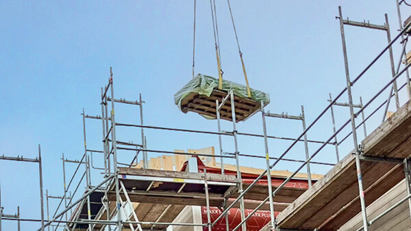Der obere Bereich einer Baustelle, auf der ein hölzernes Gebäude errichtet wird. Man sieht Holzwände und Metallgerüste. Vor dem blauen Himmel eine Palette voller Baumaterialien, mit einer Plane abgedeckt und an einem Kran hängend - den Kran selbst sieht man allerdings nicht.