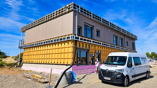 Ein neu gebautes zweistöckiges Bauwerk; in der Umgebung sind noch Bauwerkzeuge, ein Sprinter und der nackte Kiesboden mit Kabeln und Rohren. Das Gebäude selbst besteht größtenteils aus Holz; im unteren Stockwerk ist ein Gitterwerk aus Latten zu sehen, das obere ist bereits mit einer hölzernen Fassade ausgestattet. An einer Seite des Bauwerks sind außerdem Fenster und Türen zu sehen, einigermaßen unregelmäßig angeordnet.