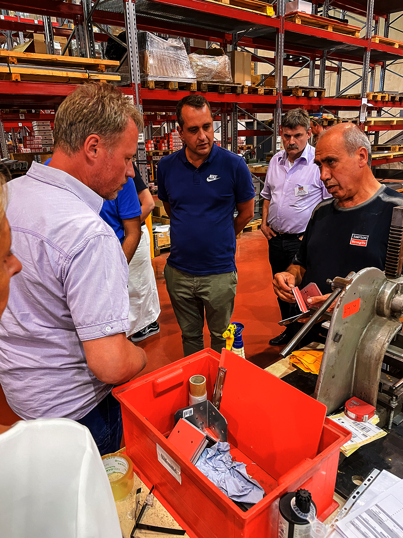 Ein Mitarbeiter von Simpson Strong-Tie erläutert den Teilnehmern im Lager die Sonderproduktion von Verbindungsmitteln anhand eines Schallschutz-Verbinders ABAI. Im Hintergrund erhebt sich ein Hochregallager.