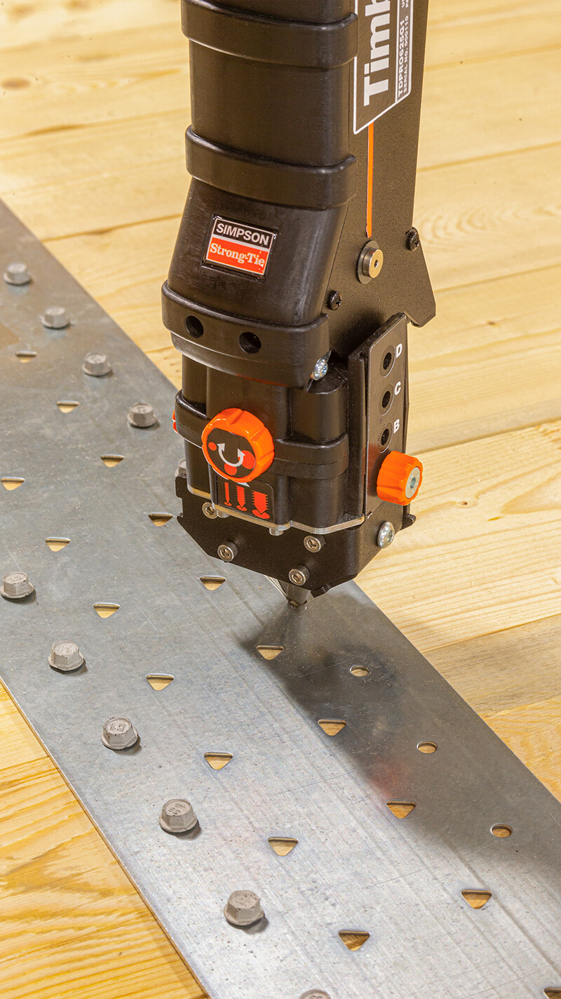 Die Spitze des Timber Drive aus der Nähe, mit der Präzisionsspitze für Holz-Metall-Verbindungen. Das Gerät dreht gerade eine Schraube in ein metallenes Lochband ein, das wiederum auf einer aus Holzbrettern bestehenden Fläche liegt.