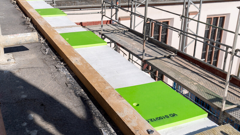 Eine Baustelle in einem höheren Stockwerk. Rechts ist das Gerüst zu sehen, im Hintergrund das Dach und die Seite eines Kirchengebäudes. Links das oberste Stockwerk des Bauwerks, an dem gearbeitet wird, und am äußersten Rand eine Reihe regelmäßig angeordneter hellgrüner Kunststoff-Polster neben einer langen hölzernen Latte.
