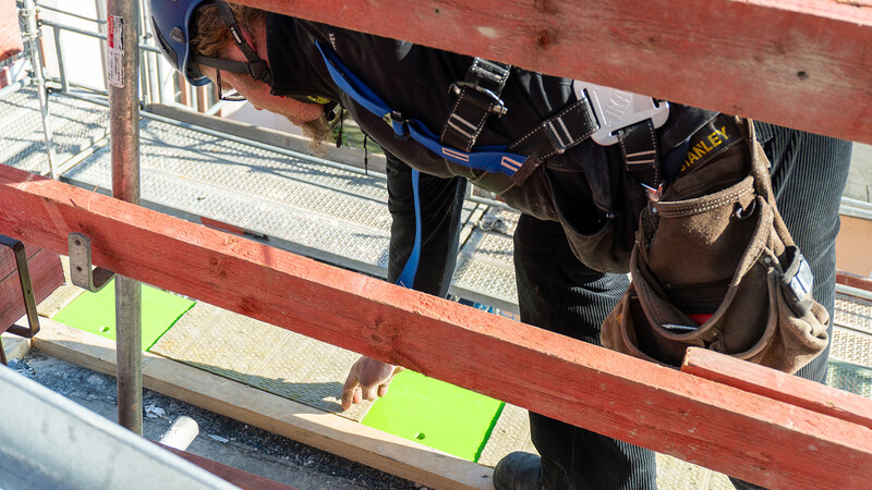 Eine Szene auf einer Baustelle, sichtbar zwischen zwei roten Holzbrettern, die als Absperrung dienen: Ein Bauarbeiter in blauer Arbeitskluft rückt ein hellgrünes Kunststoff-Polster zurecht, eines von mehreren, die bereits auf dem Boden liegen. Im Hintergrund ist das Baugerüst zu sehen, die Baustelle befindet sich offenbar in großer Höhe.