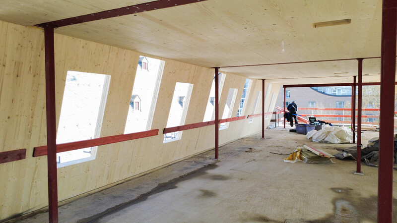 Eine Baustelle; ein neu entstehender Raum aus vorgefertigten hölzernen Elementen von Innen. Die leicht schräge Wand ist mit zahlreichen, leicht versetzten Aussparungen für die Fenster ausgestattet. Die Decke, ebenfalls aus Holz, hat nur eine einzelne quadratische Aussparung, die für eine Lüftung oder einen Kabelschacht vorgesehen sein könnte. Rötliche Bretter sind an der Wand angebracht, metallene Stützen halten die Decke. Im Hintergrund weitere Gebäude sowie ein einzelner Arbeiter in Sicherheitskluft.