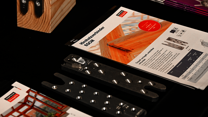Auf einer mit einer schwarzen Tischdecke bedeckten Tischplatte liegen verschiedene Objekte: die beiden Hälften eines zweiteiligen Holzverbinders, ein Produktflyer zum Holzverbinder EGCM von Simpson Strong-Tie. Oben links ist ein hölzernes Modell zu erahnen, unten links eine aufgeschlagene Broschüre.