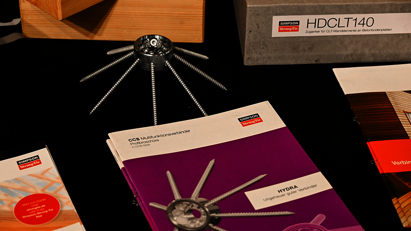 Eine mit einer schwarzen Tischdecke bedeckte Tischplatte, darauf diverse verstreute Objekte: ein Multifunktions-Holzverbinder CCS von Simpson Strong-Tie, der ein wenig wie ein metallener Seestern mit acht Zacken aussieht. Direkt darunter ist die dazugehörige Produktbroschüre in knalligem Lila. Links und rechts weitere Broschüren, oben sind zwei hölzerne Modelle zu erahnen, eines mit der Aufschrift "HDCLT140".