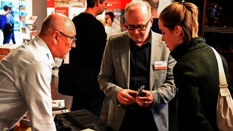 Das Bild zeigt drei Personen, die miteinander im Gespräch sind. Ein Mann hält einen zweiteiligen Holzverbinder und mustert ihn näher, ein zweiter Mann beugt sich von links heran und scheint das Teil zu erklären. Eine Frau beugt sich von rechts heran und mustert das Teil ebenfalls interessiert.