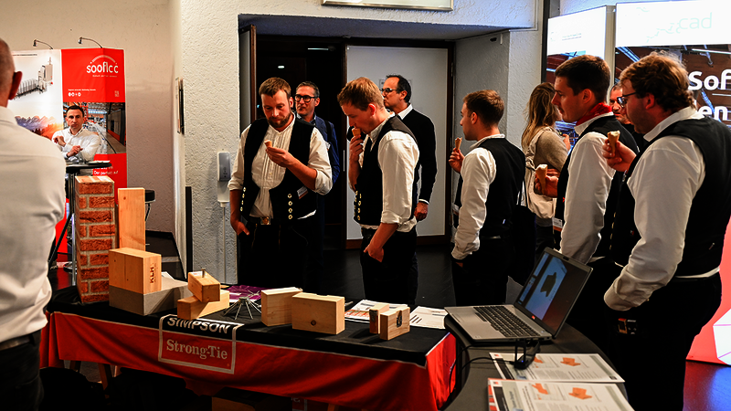 Eine Gruppe von Personen in Zimmererkluft kommt von rechts ins Bild, jeder hält eine Eistüte in der Hand. Auf den linken zwei Drittel des Bildes ist ein Tisch vom Messestand von Simpson Strong-Tie mit einer schwarz-orangeroten Tischdecke zu sehen, darauf jede Menge Broschüren, Produkte und Modelle. Die Zimmerer ganz links in der Reihe schauen nicht mehr auf ihr Eis, sondern auf das, was sich alles auf dem Tisch befindet. Hinter ihnen ist der Ausgang aus dem Raum zu sehen.