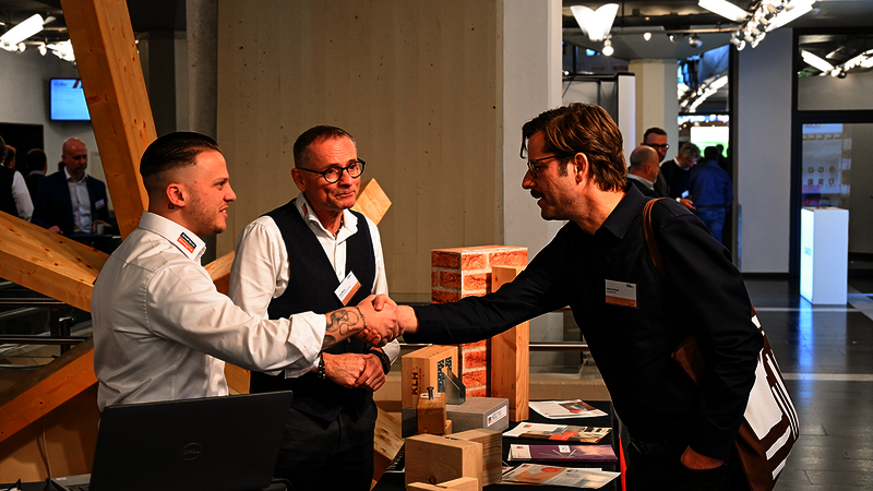 Das Bild zeigt drei Männer, von denen sich zwei über die Messetheke von Simpson Strong-Tie hinweg die Hand geben. Ein dritter Mann befindet sich ungefähr zwischen den beiden und lächelt zufrieden. Hinter dem Mann ganz links ist ein hölzernes Modell mit vielen von einem zentralen Element ausstrahlenden hölzernen Balken.