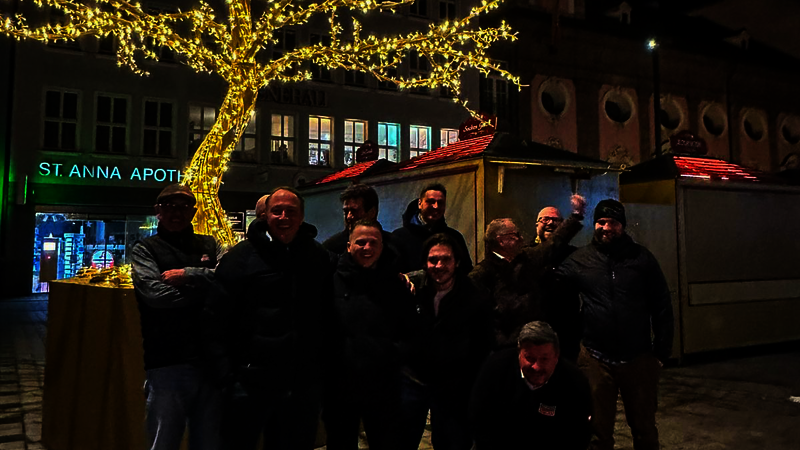 Eine Gruppe Menschen in warmen Klamotten stehen oder hocken vor einem mit weihnachtlichen Lichtern geschmückten Baum. Hinter ihnen sind Hütten eines Weihnachtsmarktes zu sehen. Das ganze Bild ist eher dunkel; es scheint Abend oder Nacht zu sein. Die Personen n der Gruppe lachen fröhlich.