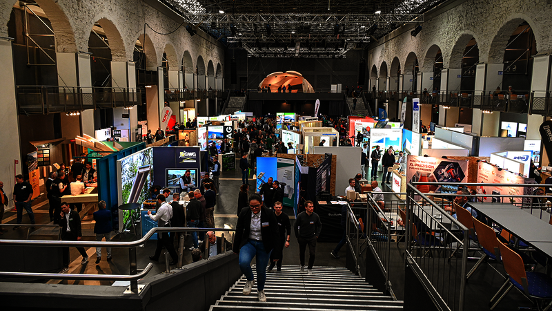 Eine Messehalle voller farbenfroher Stände und Banner, von schräg oben betrachtet. Eine Treppe führt vom Betrachter hinunter zu den Ständen. Viele Leute sind unterwegs. Links und rechts tragen große Steinbögen die Decke; es scheint sich um ein historisches Gebäude zu handeln. 
