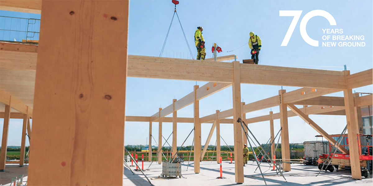 Eine Holzbaustelle von leicht unten betrachtet. Auf einem langen Querbalken stehen zwei Bauarbeiter oder Zimmerleute in hellgelben Sicherheitsklamotten, Blaumännern und Sicherheitshelmen. Hinter ihnen hebt ein Kran weitere Balken an. Der Boden der Baustelle besteht aus Beton. Im rechten oberen Bildrand eine große 70 mit dem Text "YEARS OF BREAKING NEW GROUND".